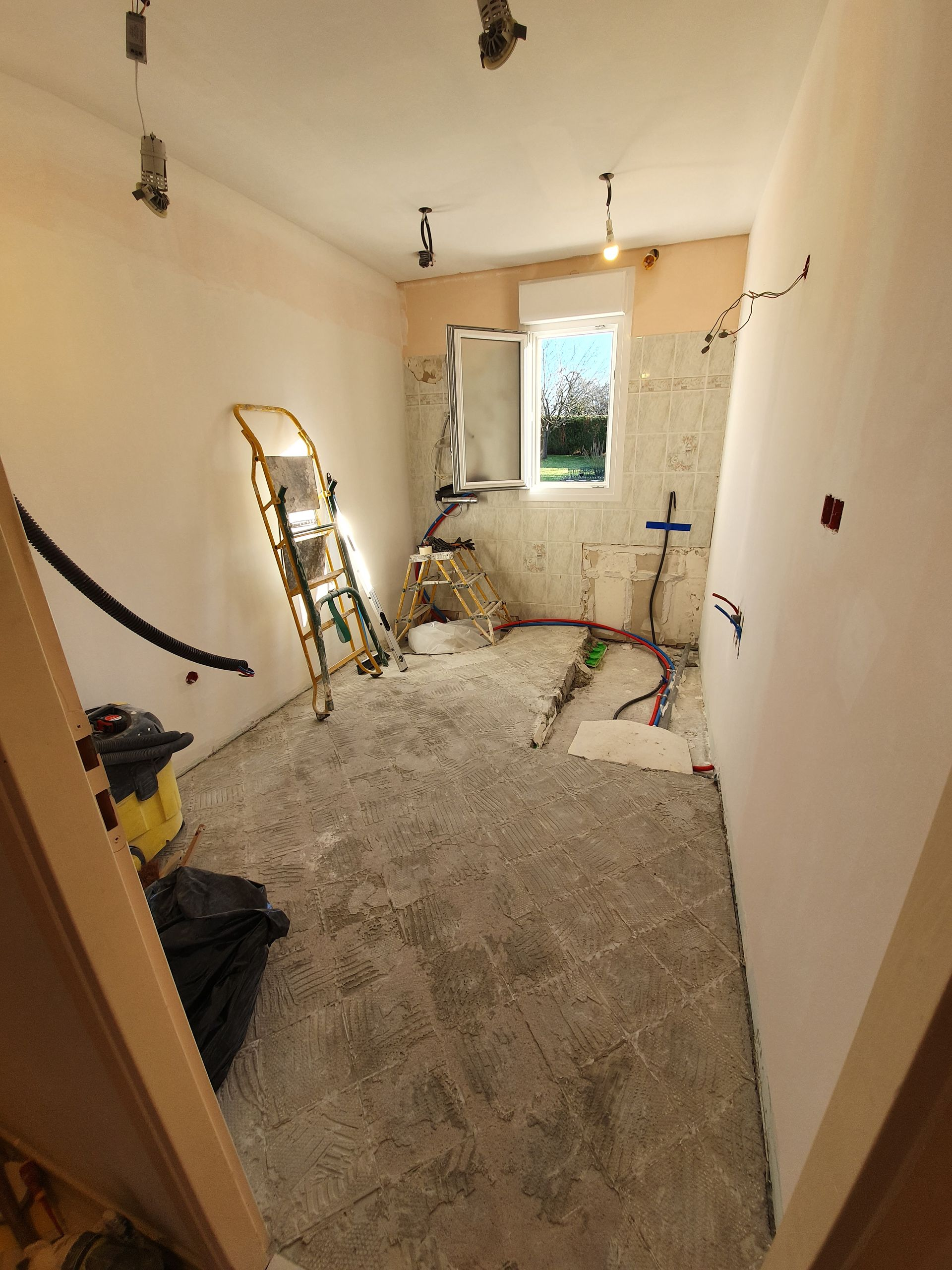 Rénovation de la pièce : mur à moitié fini et débris, l’autre côté est peint en blanc avec un parquet. La lumière entre par la fenêtre.