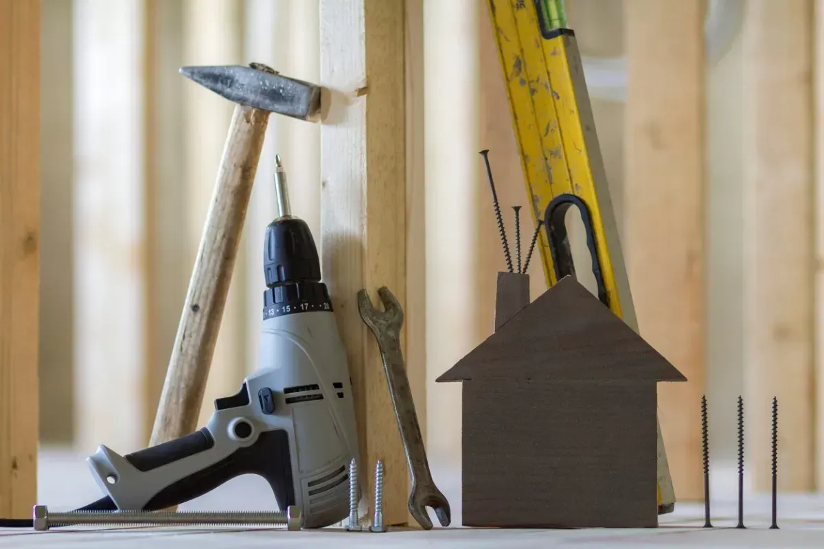 Herramientas de construcción, incluido martillo, taladro, nivel y tornillos, junto a un pequeño recorte de una casa.