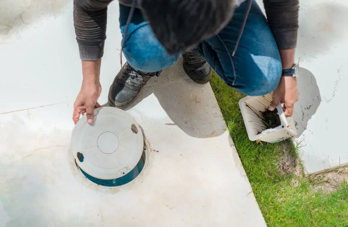 Persona arrodillada junto a una estructura blanca, quitando una tapa blanca, al lado de césped verde.