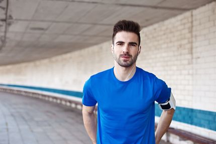 Homem de camisa azul, braçadeira e fones de ouvido está parado embaixo de um viaduto de concreto curvo.