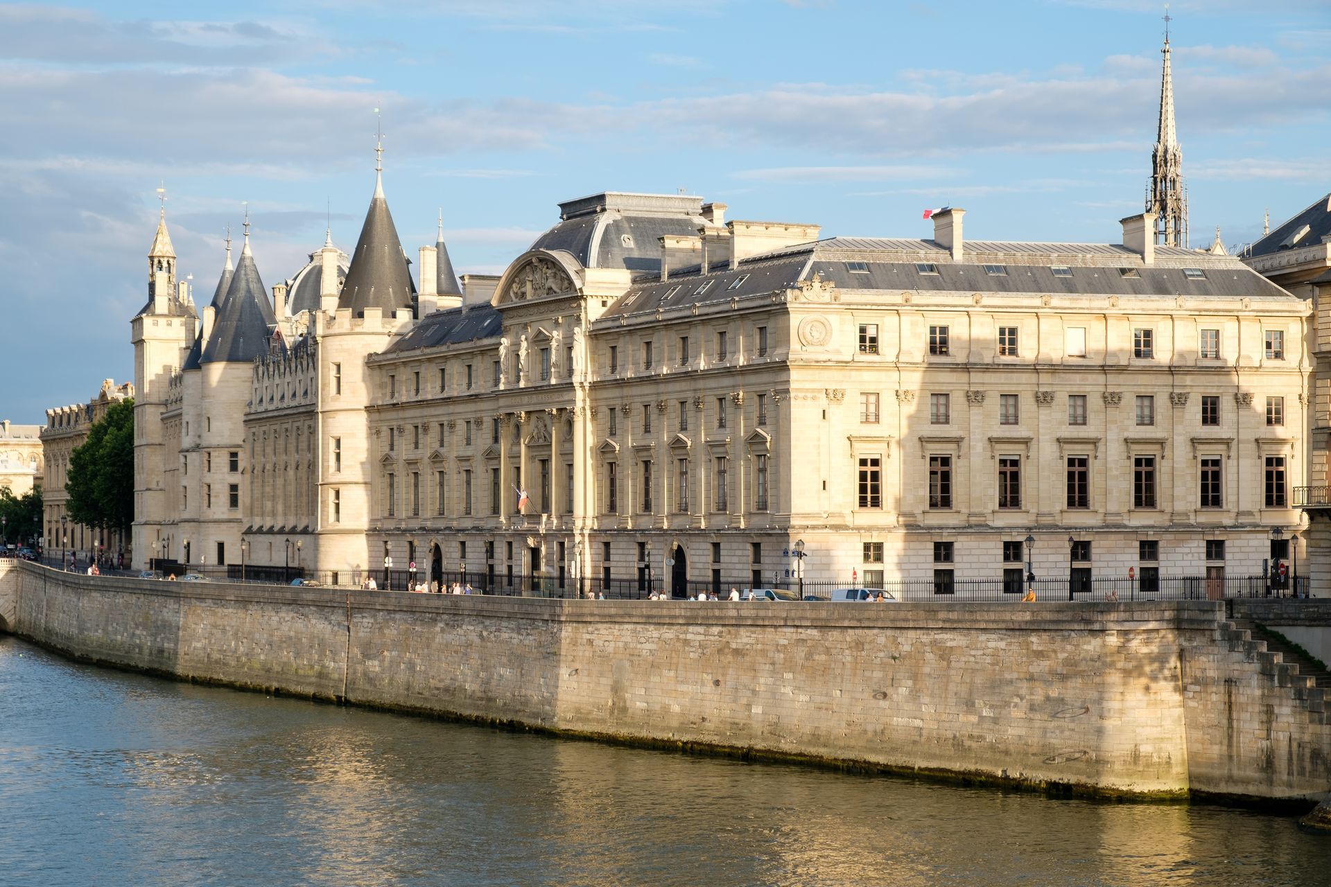 Bâtiments historiques en pierre le long d'une rive sous un ciel bleu, avec des tours et un pont sur la gauche.