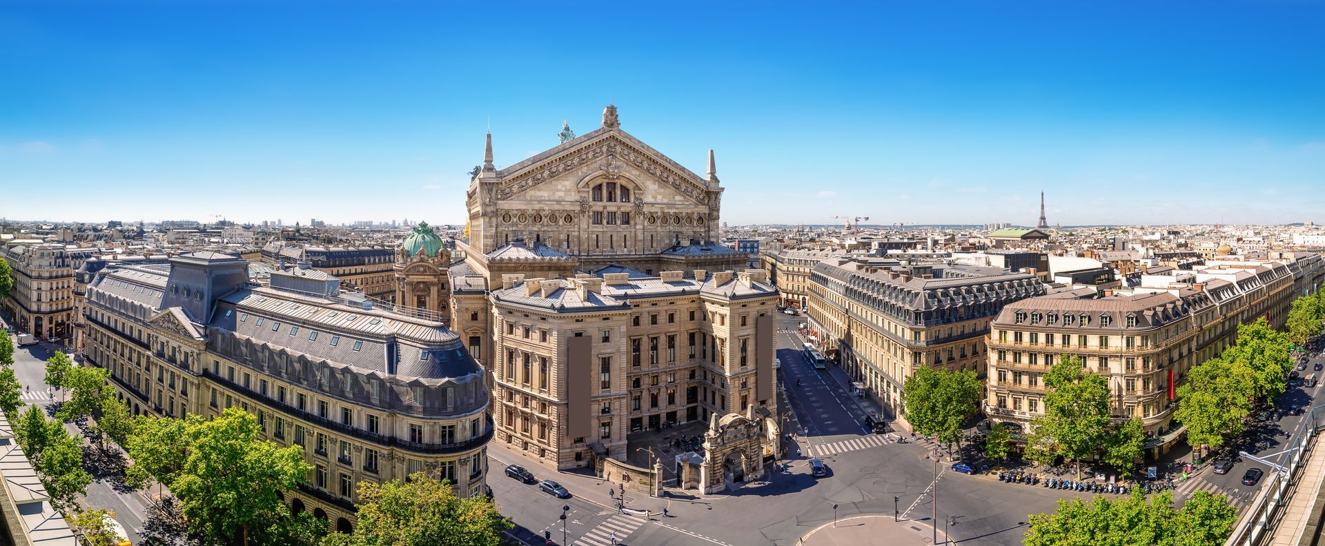 Vue aérienne panoramique d'un grand bâtiment historique sur une place animée d'une ville européenne, sous un ciel dégagé.