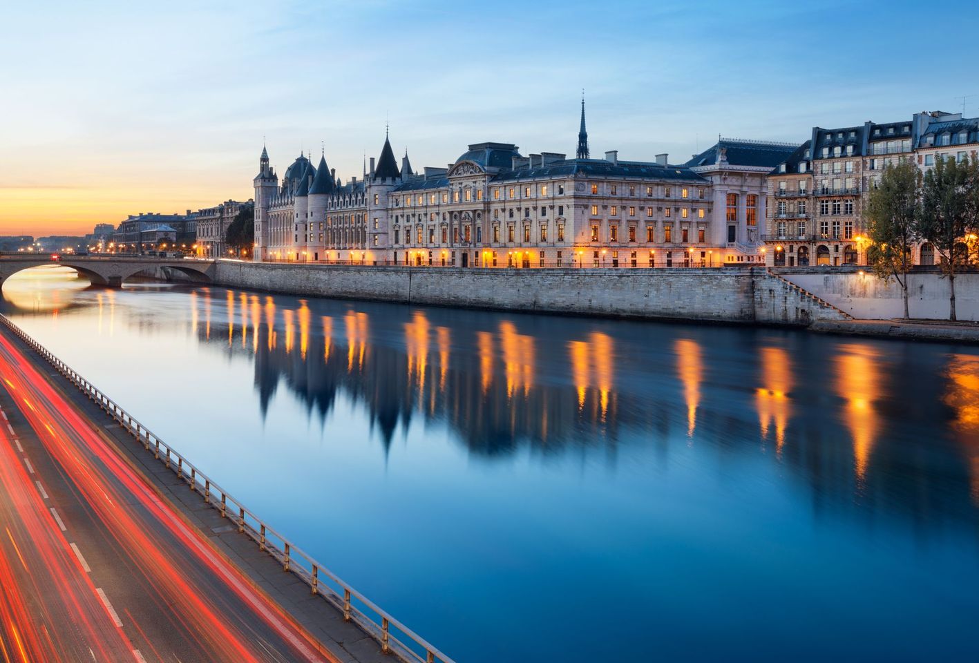 Coucher de soleil sur une ville riveraine, avec ses bâtiments illuminés et le reflet de ses ponts sur l'eau calme.
