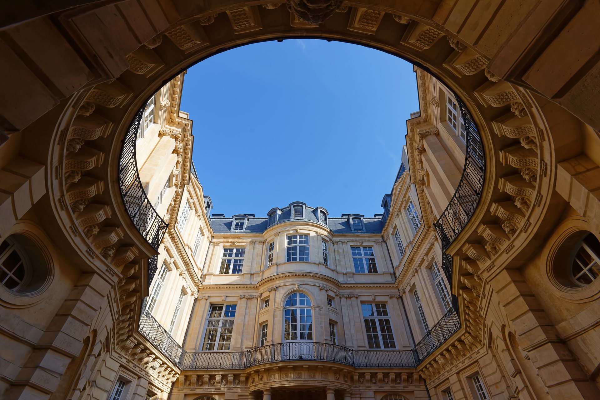 En levant les yeux à travers l'arche en pierre d'une cour intérieure, on aperçoit des bâtiments ornés de couleur crème et un ciel bleu.