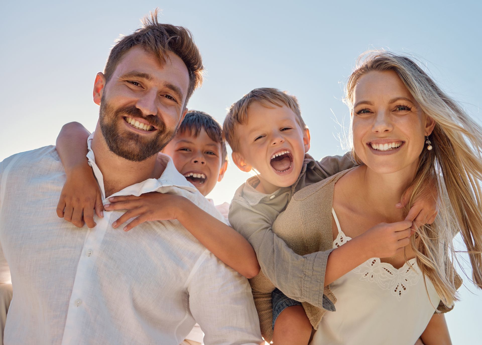 Une famille de quatre personnes, souriante, pose ensemble à l'extérieur sous un soleil radieux, avec deux enfants sur le dos de leurs parents.