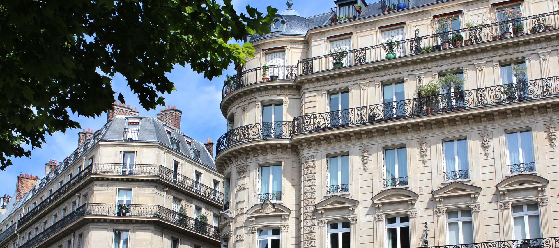 Élégants bâtiments européens couleur crème, aux balcons ornés, se détachant sur un ciel bleu, encadrés par les arbres.