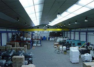 Interior del almacén con mercancía empaquetada, maquinaria y grúa puente. Claraboyas y luces fluorescentes iluminan el espacio.