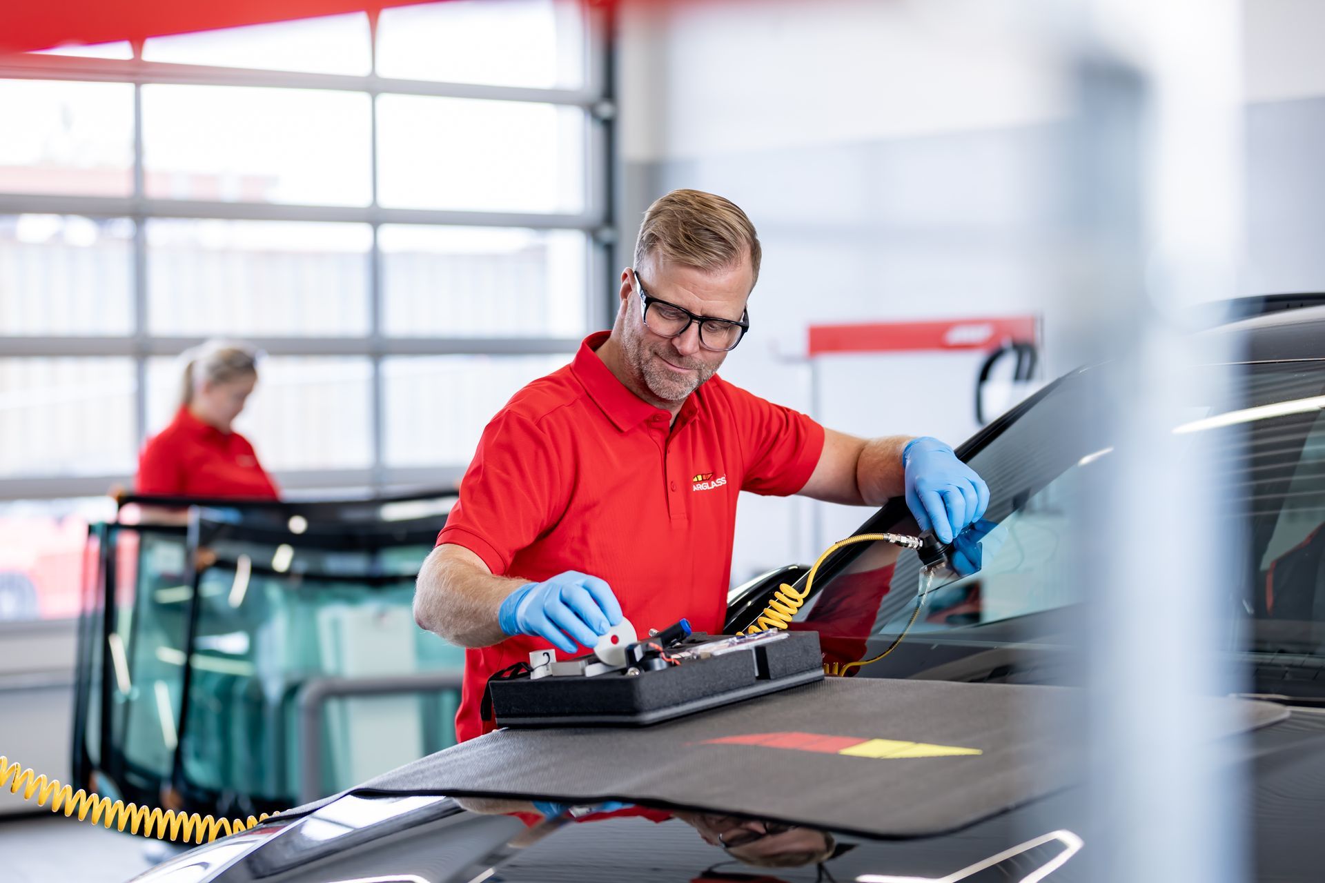 Maður í rauðri skyrtu gerir við framrúðu bíls í verkstæði. Hann er með hanska og öryggisgleraugu.