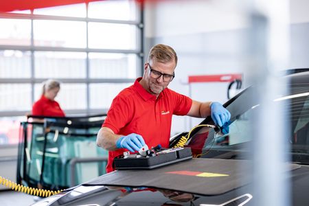 Maður í rauðri skyrtu gerir við framrúðu bíls í verkstæði. Hann er með hanska og öryggisgleraugu.