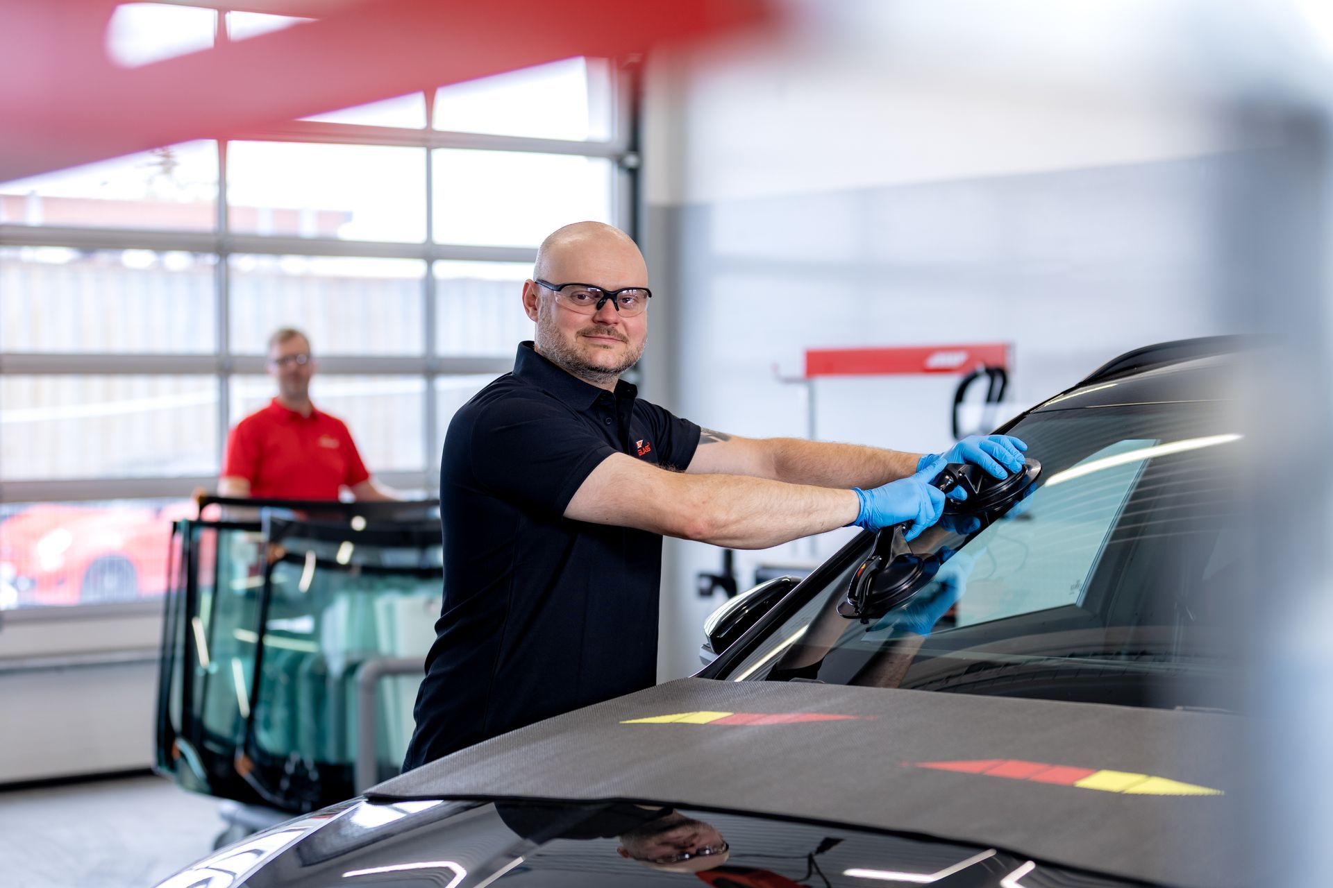 Vélvirki setur upp framrúðu á bíl, klæddur í hanska, í bílskúr. Annar vélvirki stendur þar nærri.