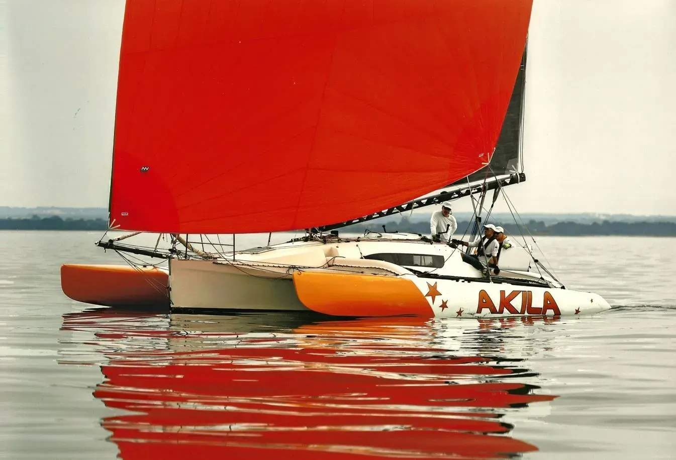 Un trimaran blanc aux coques orange vif et à la grande voile rouge glisse sur une eau calme, avec deux personnes à bord.