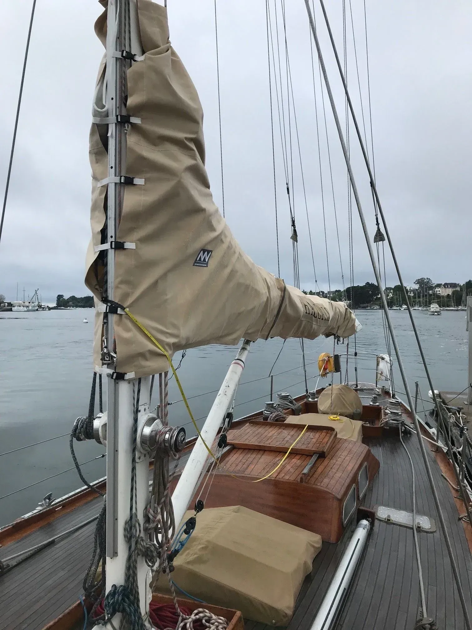 Le pont d'un voilier, avec un mât recouvert de beige et une voile enroulée, agrémenté d'éléments en bois et de gréement, sur une voie navigable calme.