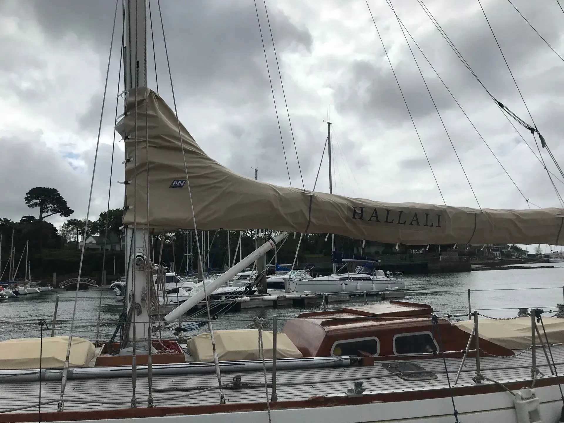 Une housse de voile beige marquée HALLALI sur la bôme d'un voilier amarré dans un port sous un ciel nuageux.