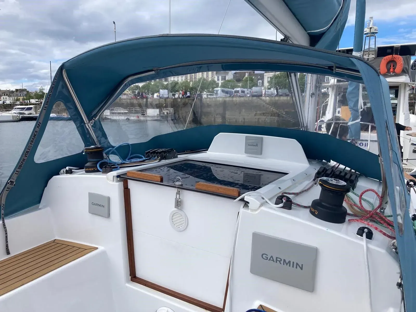 Le cockpit d'un voilier avec un pare-brise en toile bleue, une console blanche avec des écrans Garmin et des winchs noirs.