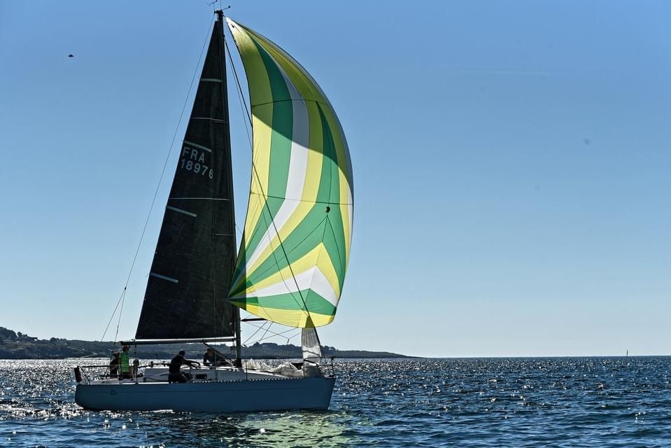 Un voilier à grand-voile sombre et spinnaker vert, jaune et blanc naviguant sur un océan calme et ensoleillé.