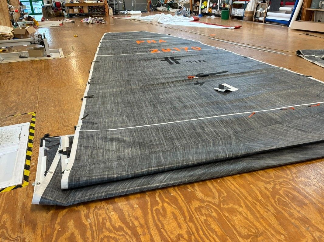 Une personne vêtue d'une veste bleue dirige un voilier vert clair à pont en bois sur une eau agitée, sous une voile d'un blanc éclatant.