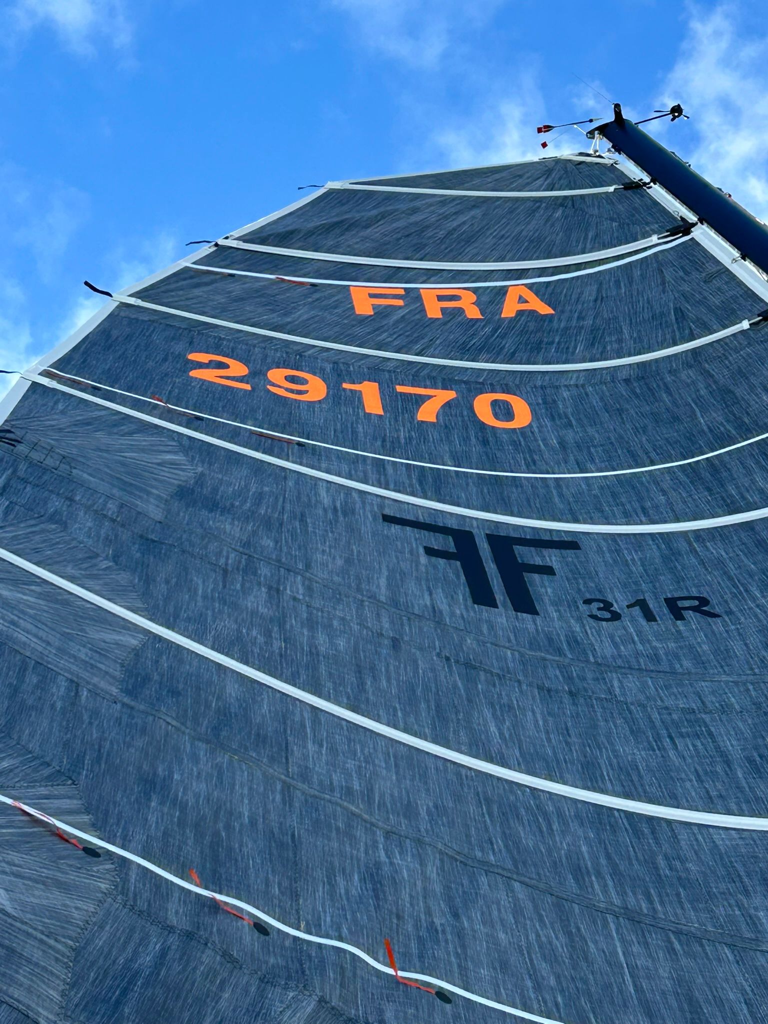 Un voilier gîte près d'une côte rocheuse tandis que trois personnes sont assises sur le pont, toutes vêtues de vêtements de plein air.