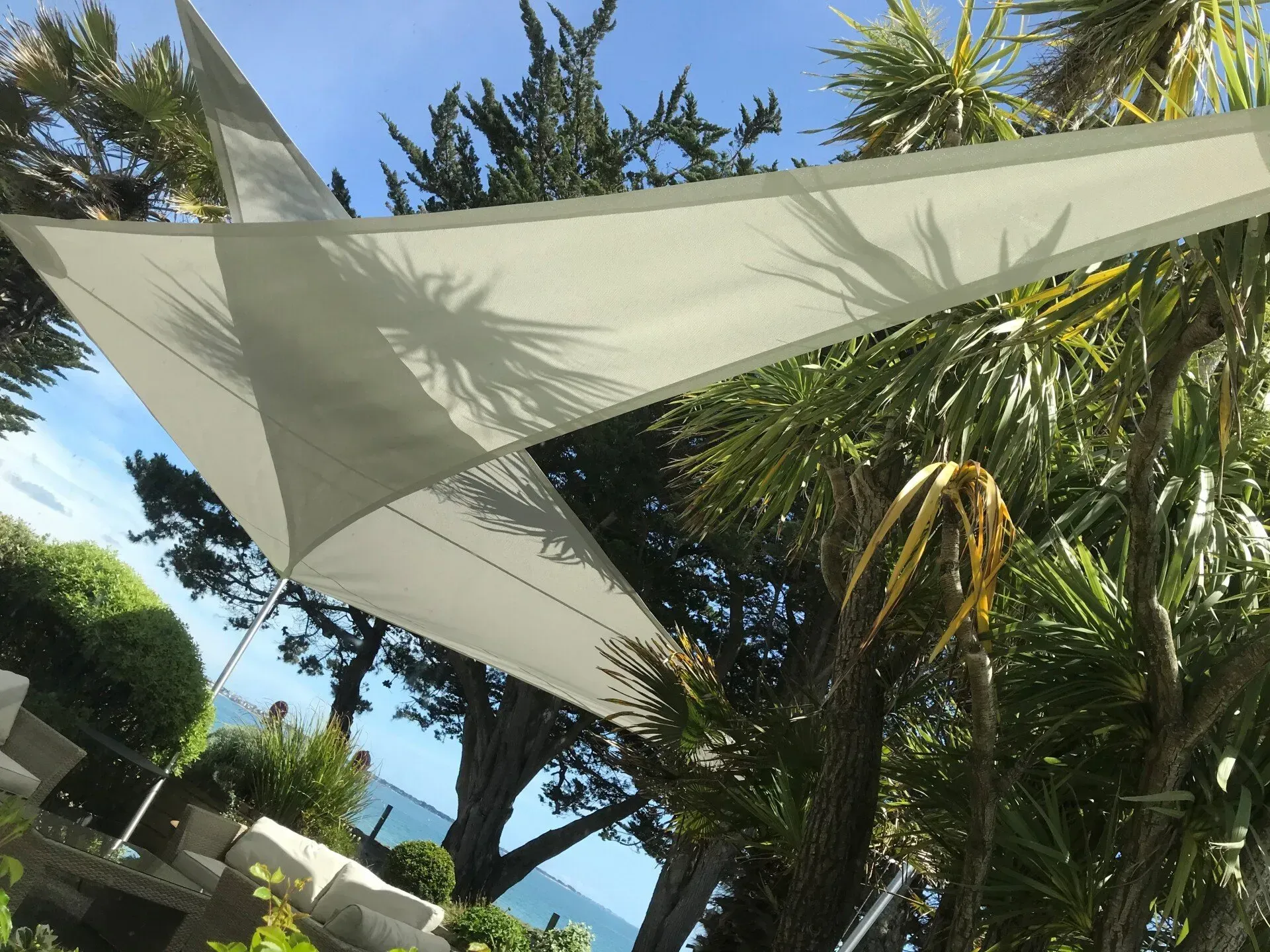 Une voile d'ombrage triangulaire blanche était tendue au-dessus d'une terrasse de jardin ensoleillée, agrémentée de palmiers et offrant une vue partielle sur la mer.