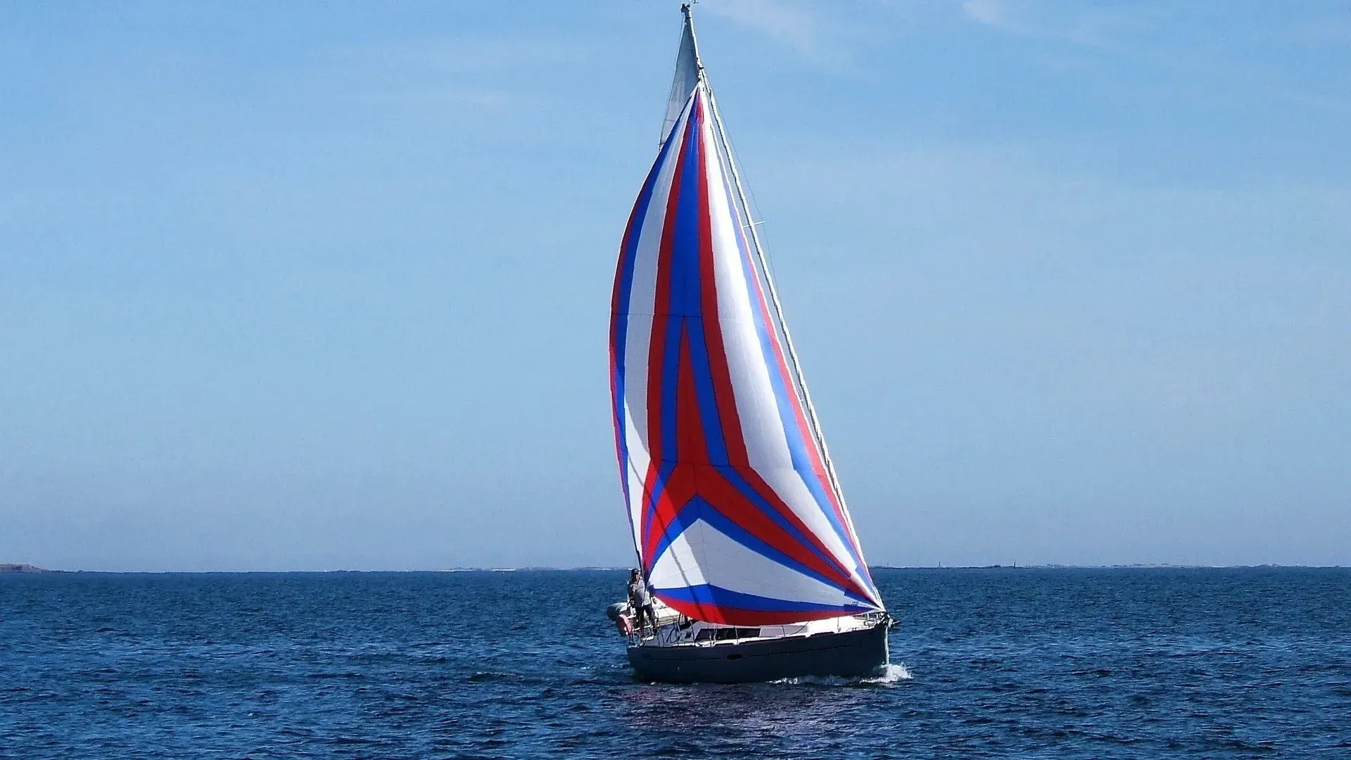 Un voilier arborant une grande voile spinnaker rouge, blanche et bleue aux couleurs éclatantes naviguant sur une eau bleue et dégagée sous un ciel clair.
