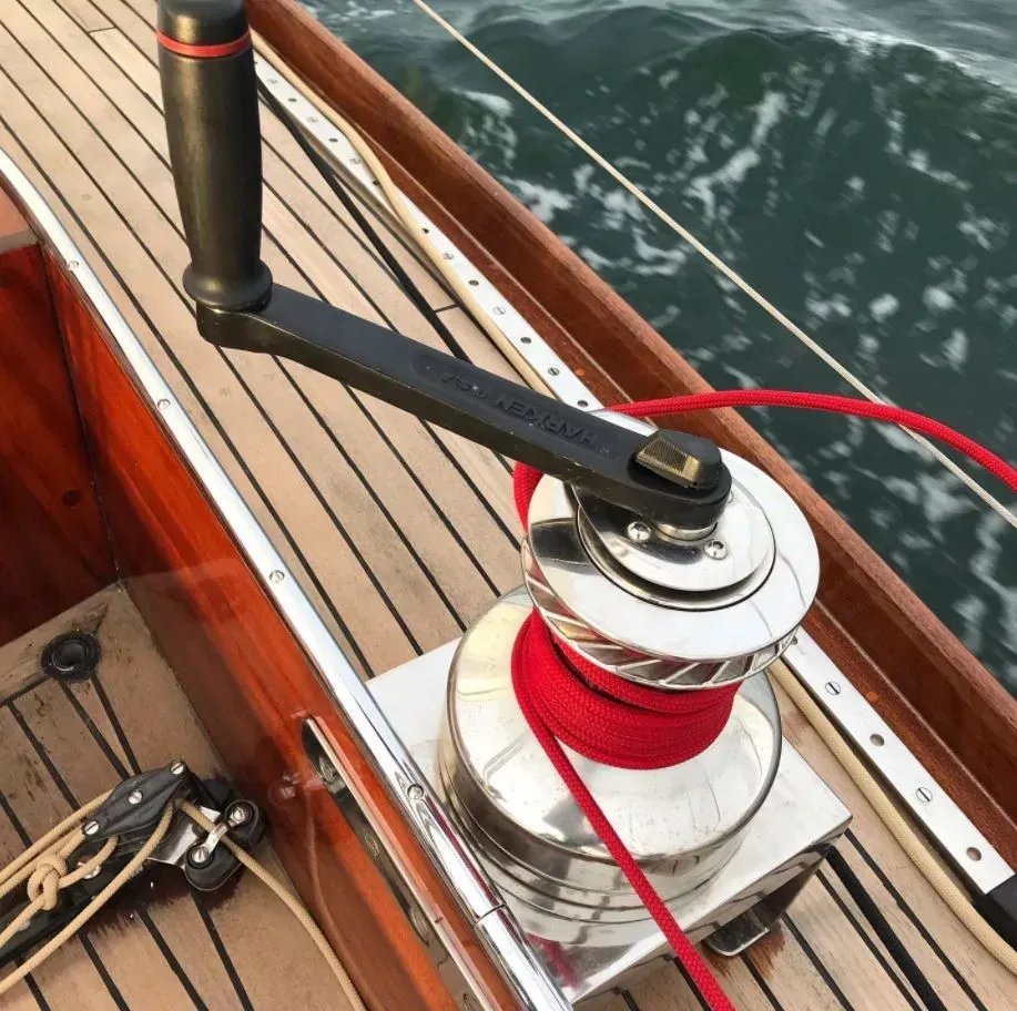 Un treuil de voilier chromé avec une poignée noire, fixé par une corde rouge sur un pont en bois, face à l'eau.