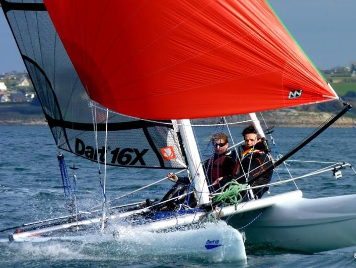 Deux personnes naviguant sur un catamaran Dart 16X, avec une grande voile orange, un ciel bleu clair et la côte en arrière-plan.