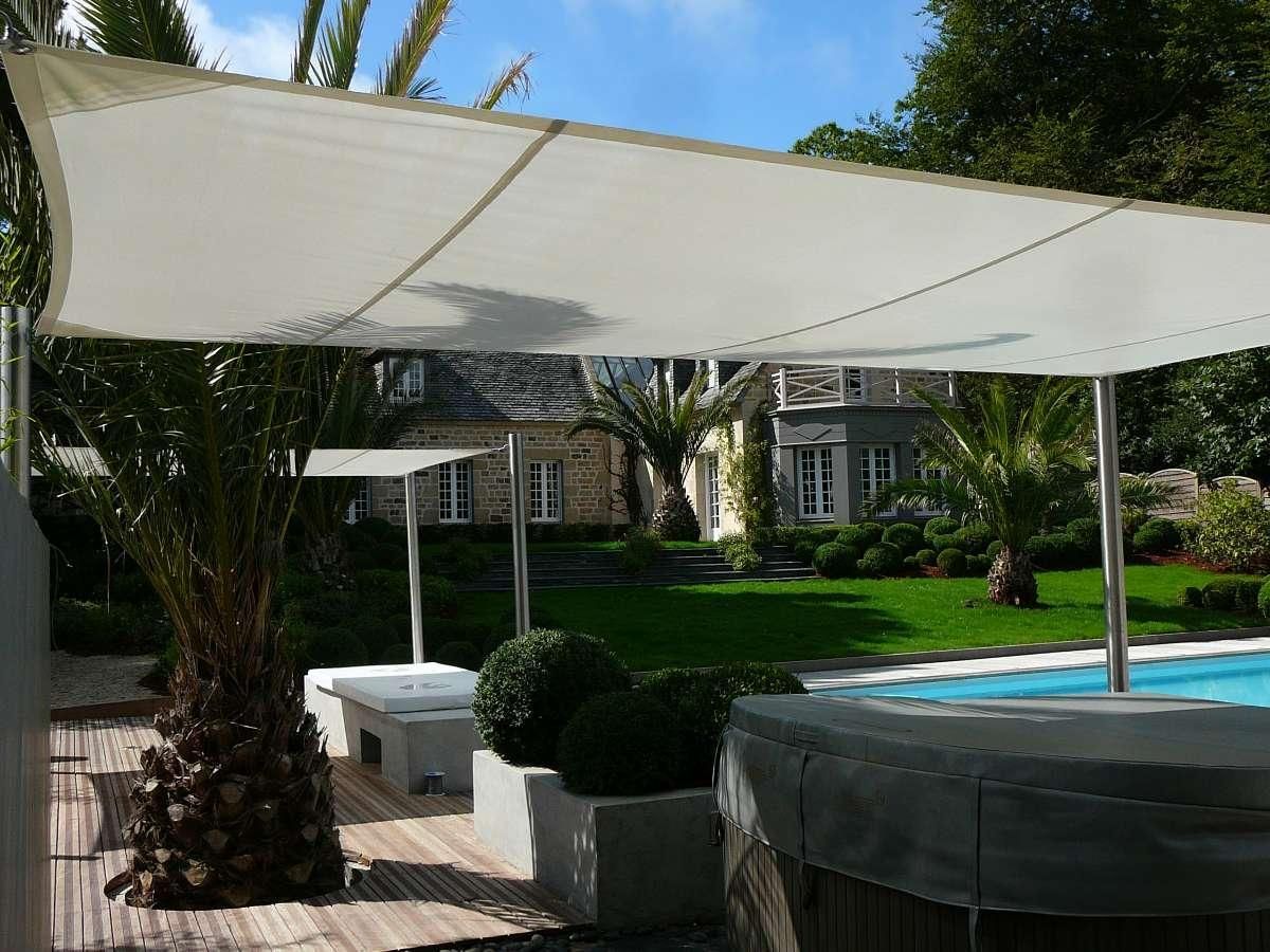 Une voile d'ombrage rectangulaire blanche recouvre une terrasse en bois agrémentée d'un palmier, de jardinières en pierre et d'une piscine dans le jardin.