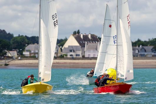 Deux voiliers, l'un jaune et l'autre rouge, filent à toute allure sur l'eau près d'une plage de sable et de bâtiments, sous un ciel nuageux.
