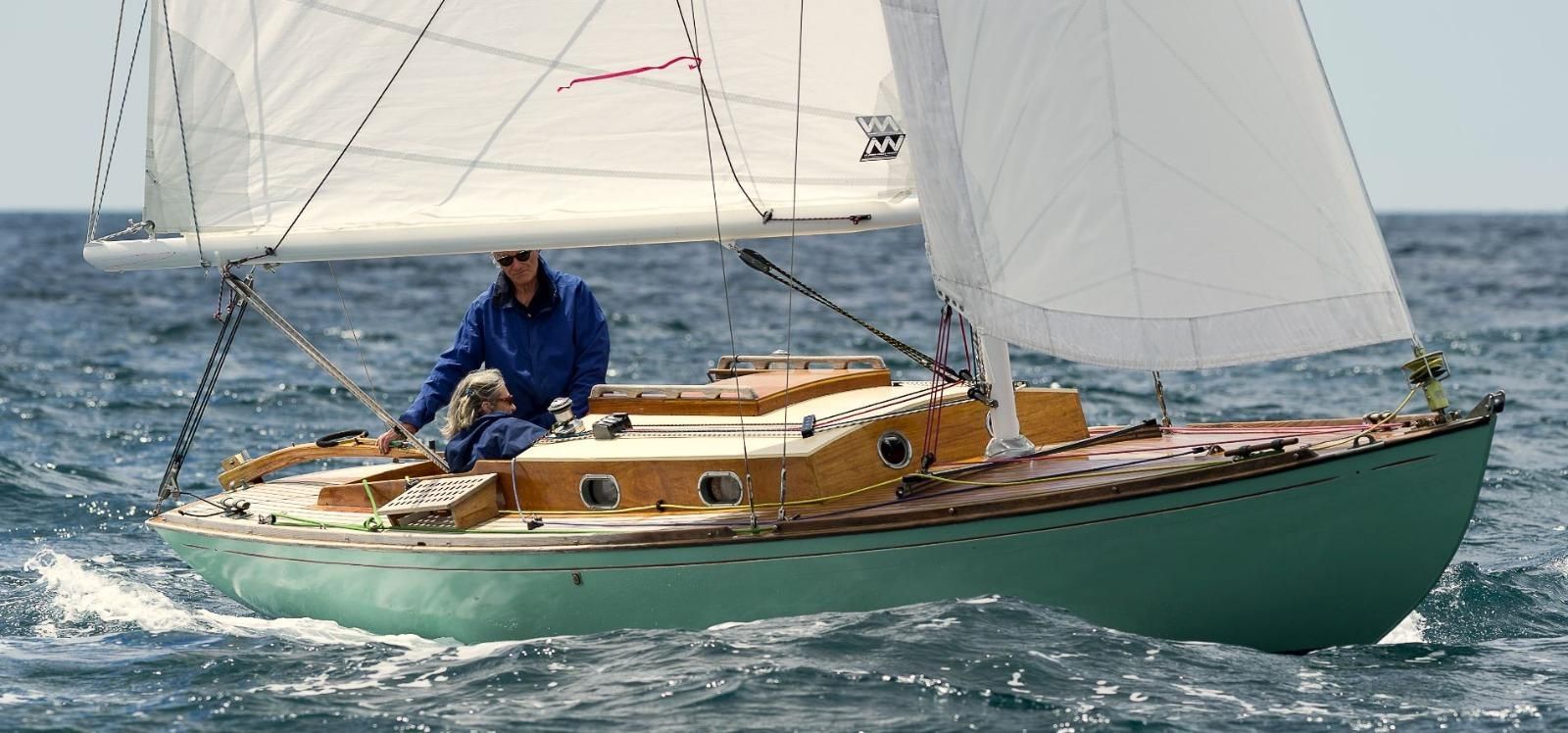 Une personne vêtue d'une veste bleue dirige un petit voilier à la coque vert menthe et au pont en bois sur une eau bleue agitée.