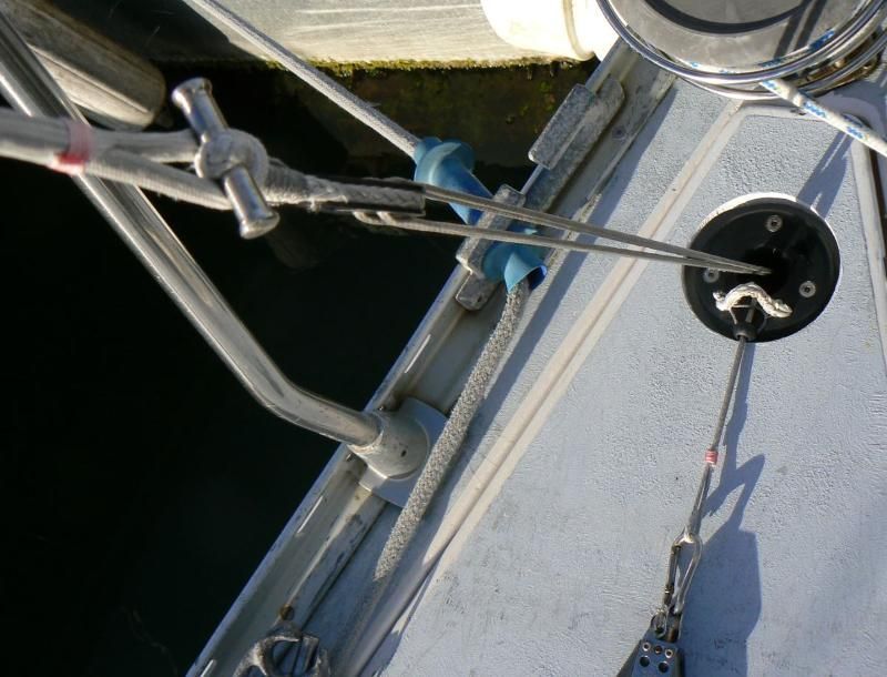 Gros plan sur le pont d'un voilier montrant le gréement métallique, un guide en plastique bleu et un système de poulies fixé à un support noir.