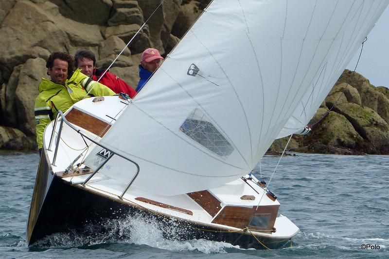 Trois personnes naviguant sur un petit bateau à grande voile blanche près de falaises rocheuses au bord de l'eau.