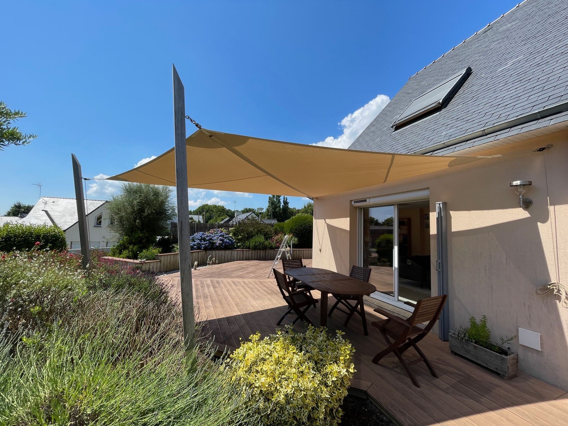Une voile d'ombrage beige protège une terrasse en bois avec une table et des chaises, à côté d'une maison sous un ciel bleu azur.