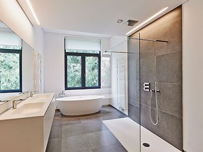 Baño moderno con azulejos grises, bañera blanca, ducha de cristal y ventana grande.