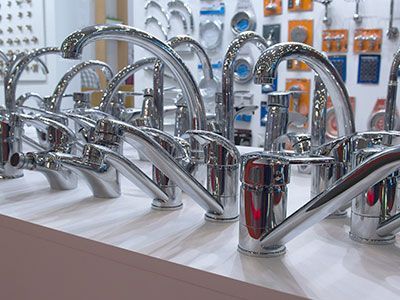 Grifos de baño con acabado cromado exhibidos en un mostrador, con más grifos y accesorios de plomería en el fondo.