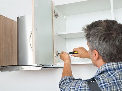 Una persona con una camisa a cuadros está instalando una puerta de armario con un destornillador; entorno de cocina.