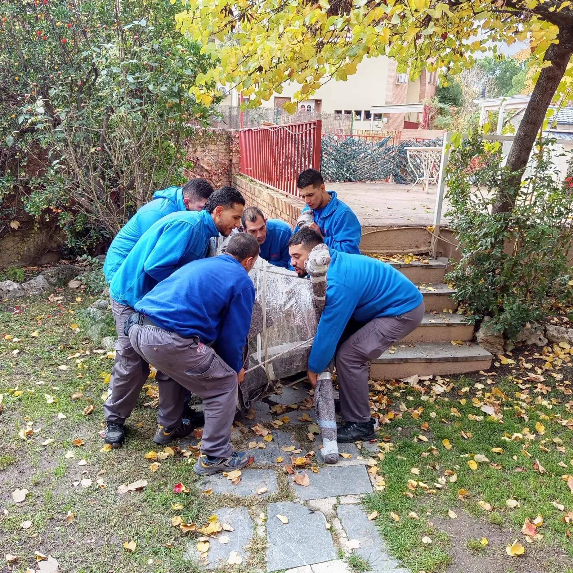Seis personas vestidas con camisas azules y pantalones grises levantan un objeto de piedra al aire libre.