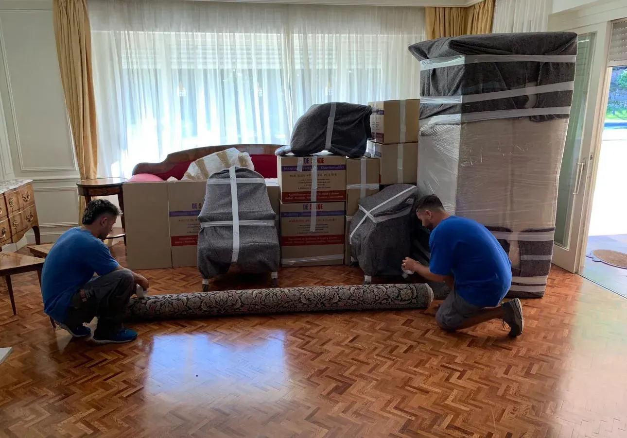 Dos operarios de mudanzas preparan una alfombra enrollada. Cajas y muebles envueltos están apilados