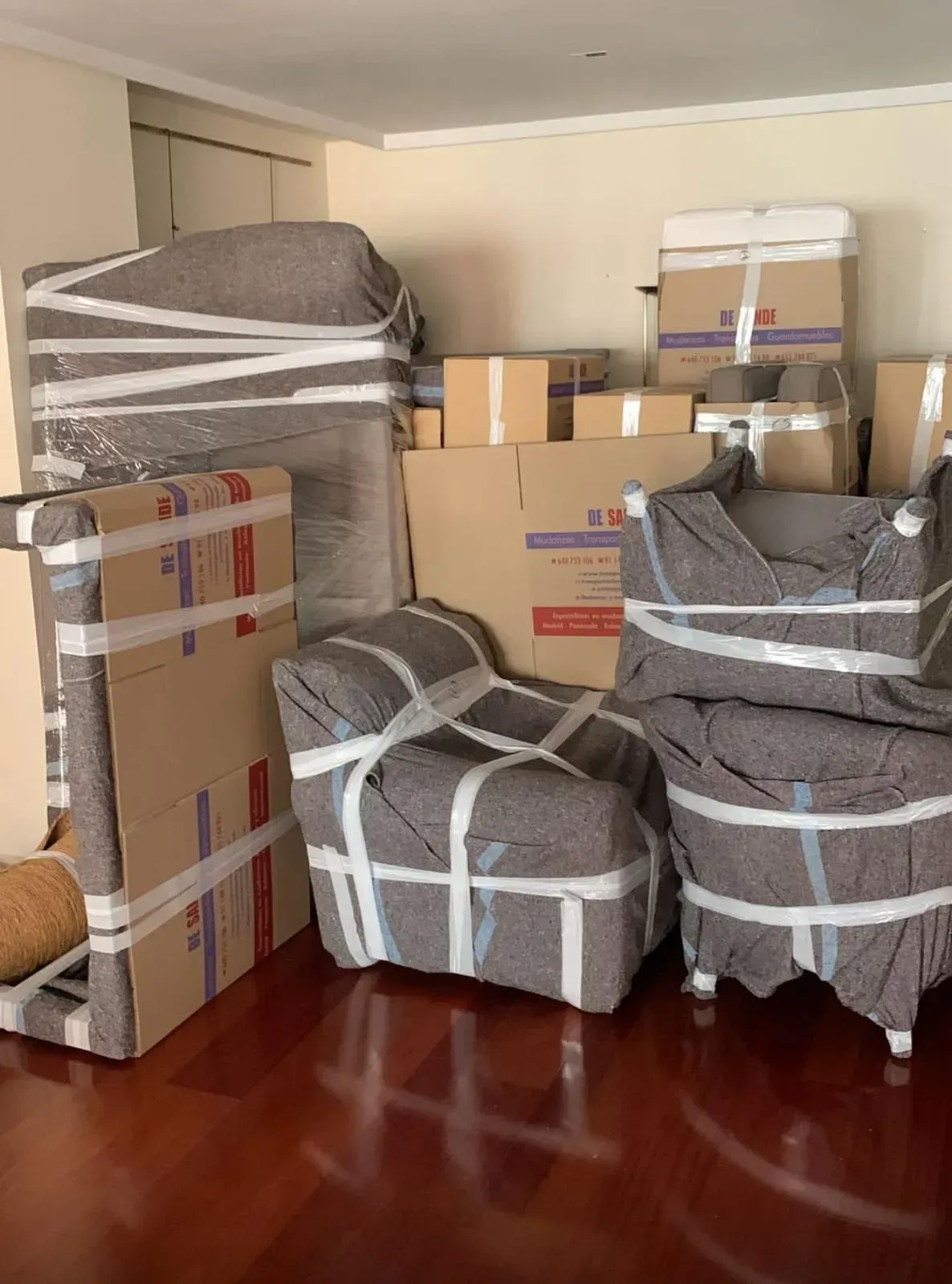 Cajas de cartón apiladas en una habitación, listas para ser trasladadas o almacenadas, con una ventana y una mesa a la vista.