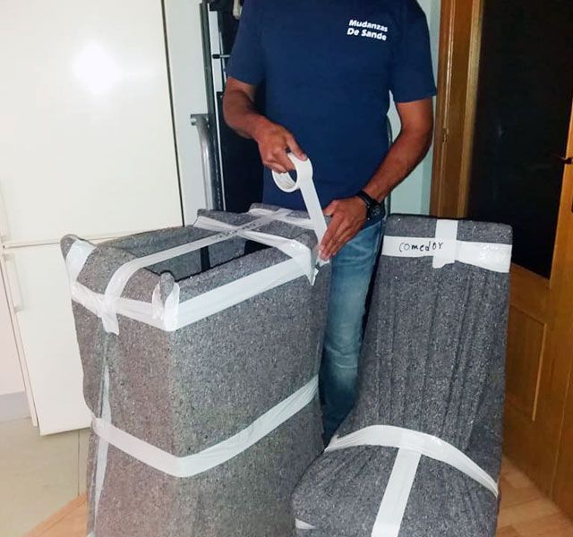 Persona que está pegando con cinta adhesiva un mueble y una silla envueltos, presumiblemente preparándose para una mudanza.