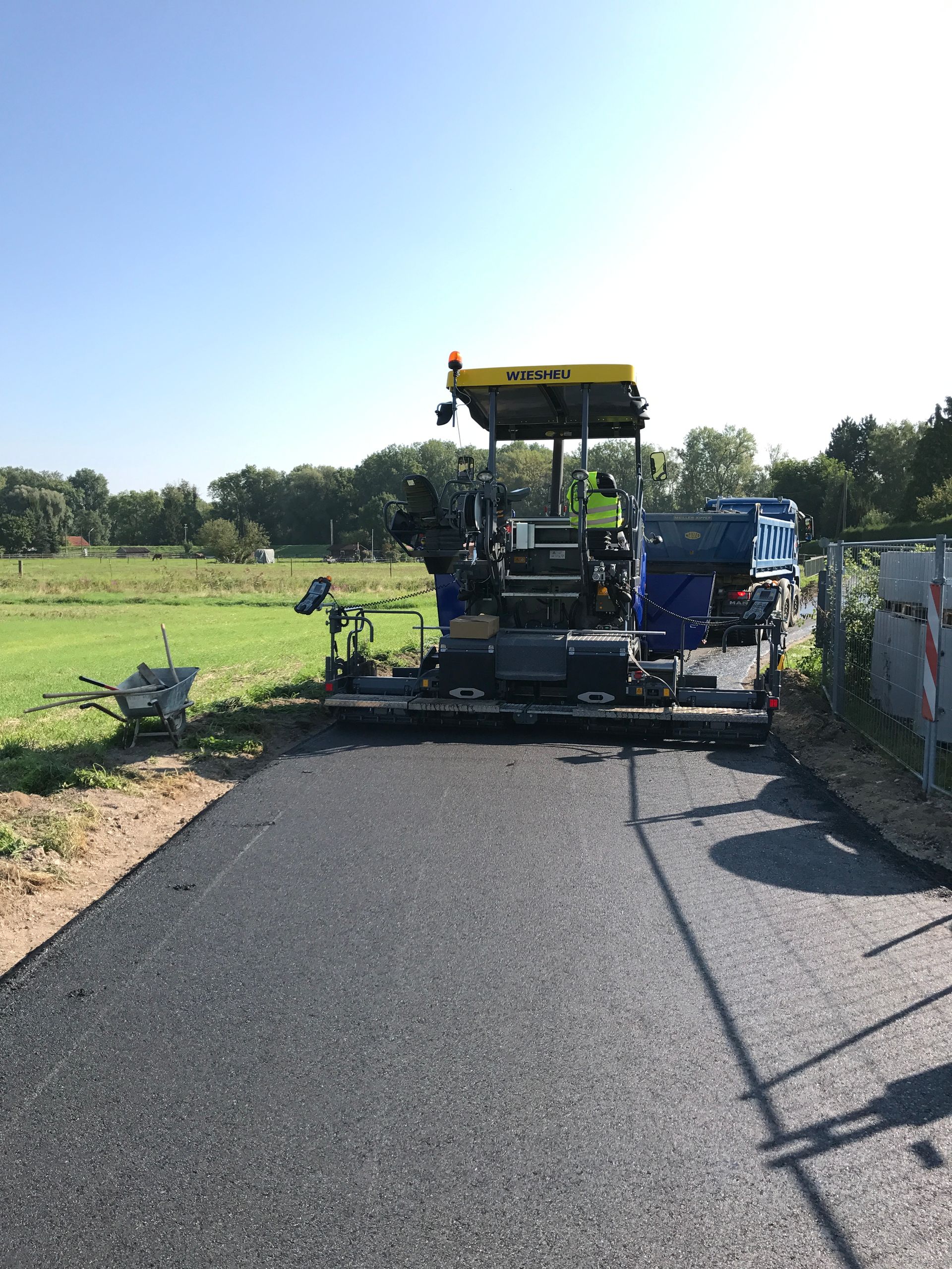 Ein Straßenarbeiter in orangefarbener Schutzkleidung verteilt Asphalt mit einer Schaufel; im Hintergrund sind Baufahrzeuge zu sehen.