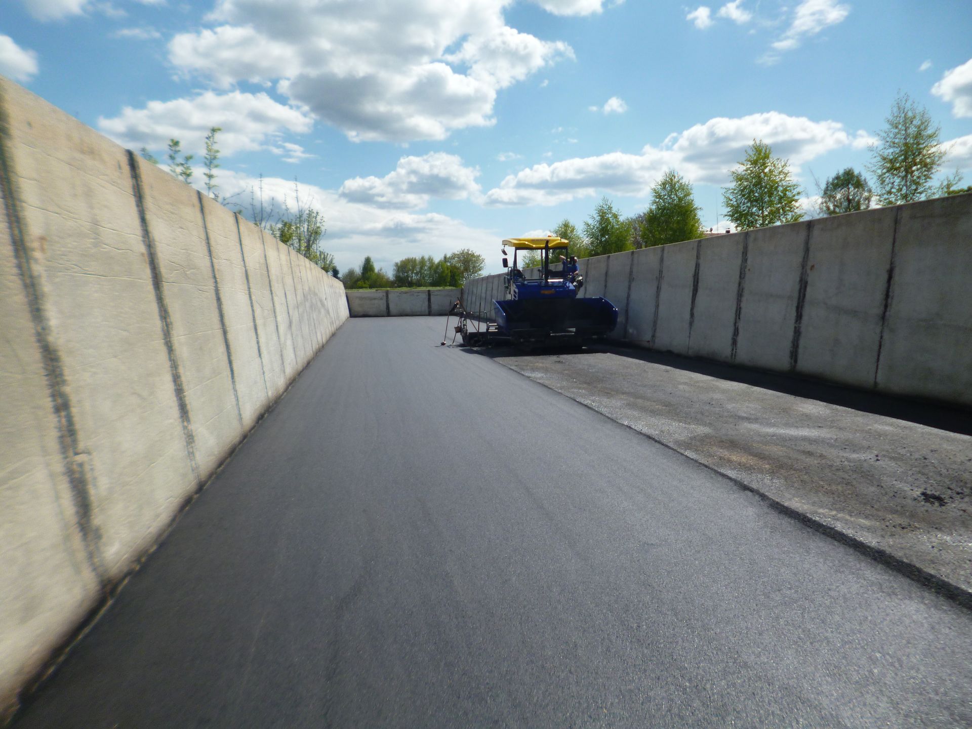 Ein weißer Lastwagen fährt auf einem Feldweg neben einem langen, weißen Getreidesack auf einem grünen Feld unter blauem Himmel.