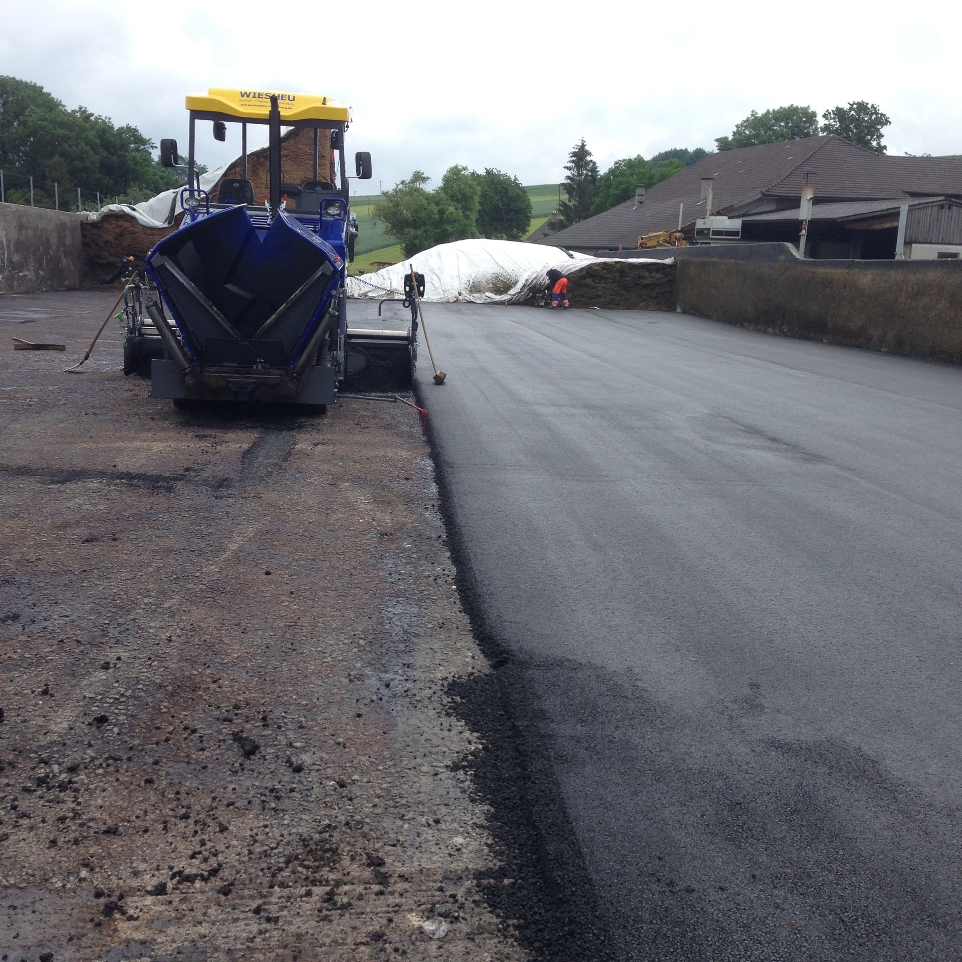 Ein weißer Lastwagen fährt auf einem Feldweg neben einem langen, weißen Getreidesack auf einem grünen Feld unter blauem Himmel.