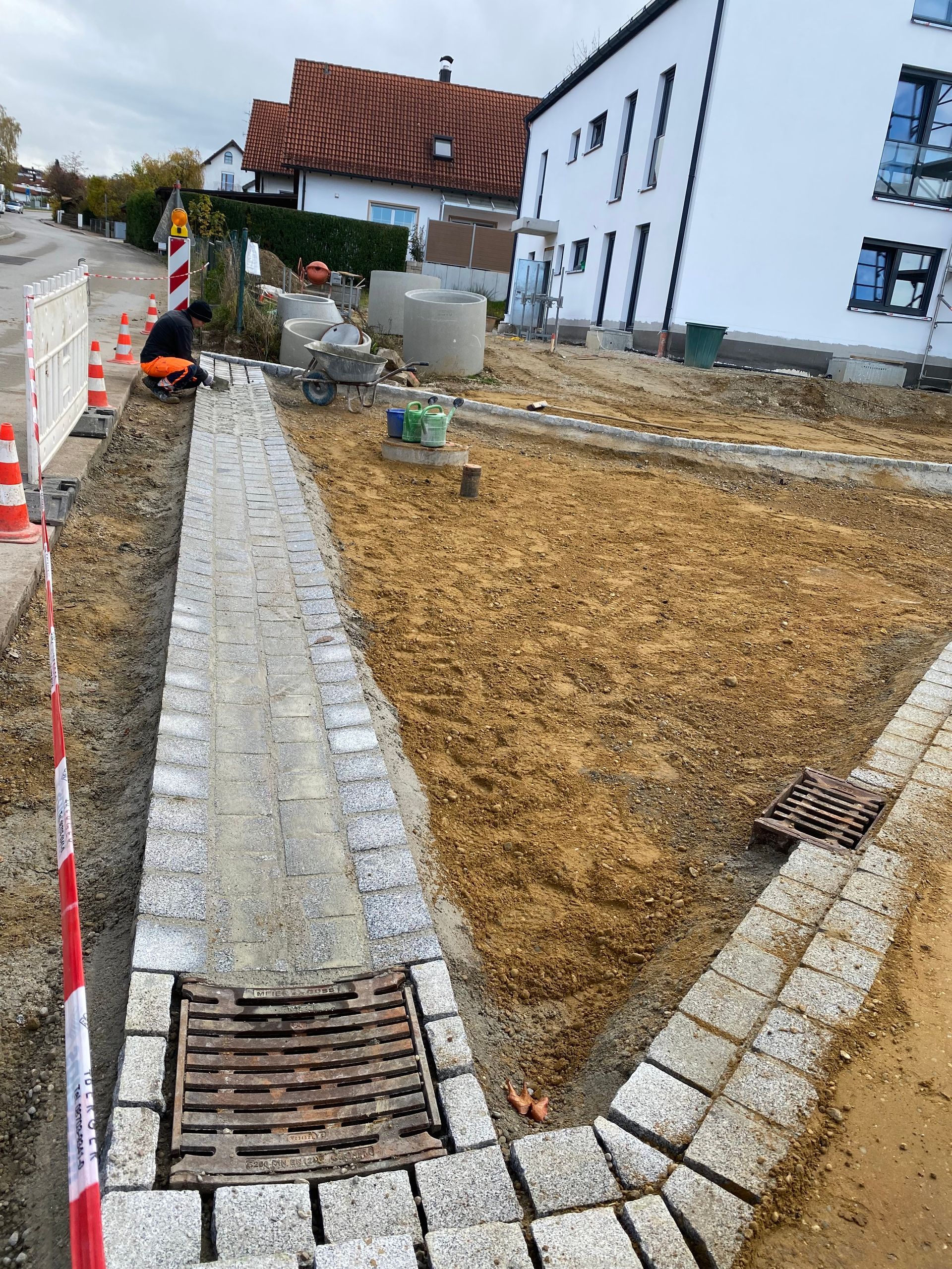 Eine Person verlegt kniend graue Pflastersteine auf einer sandigen Außenfläche. Sie trägt Handschuhe.