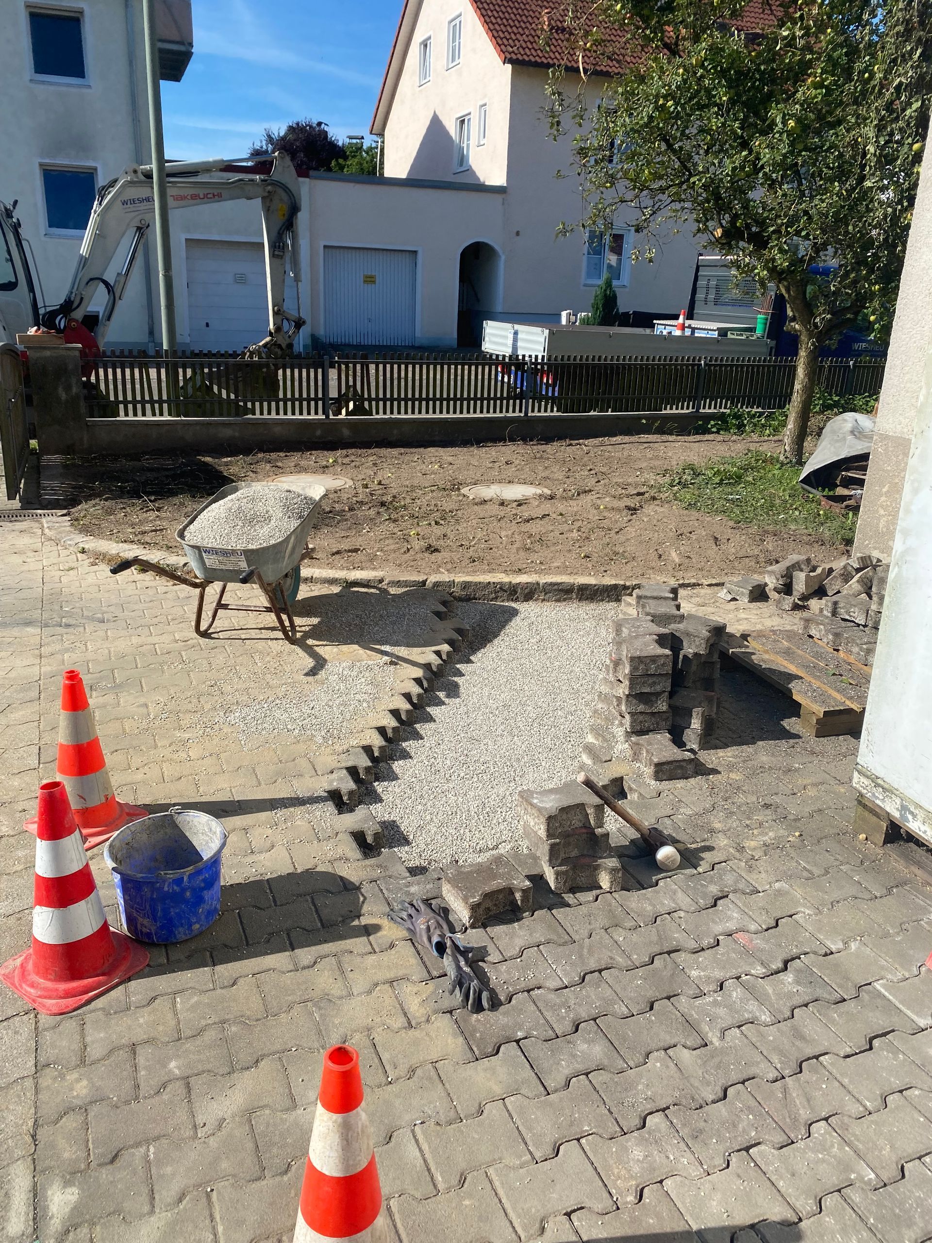 Eine Person verlegt kniend graue Pflastersteine auf einer sandigen Außenfläche. Sie trägt Handschuhe.