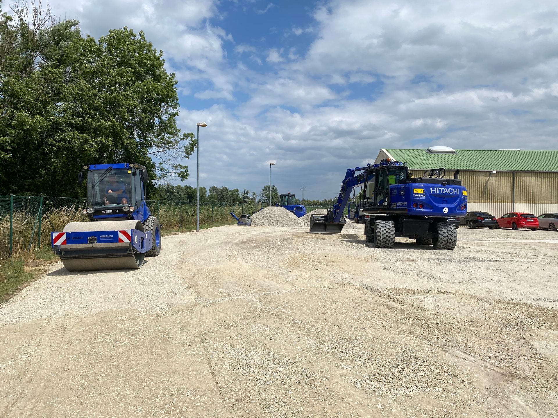 Ein grüner Baggerlader schaufelt Erde auf einer Baustelle, im Hintergrund Bäume, bewölkter Himmel.