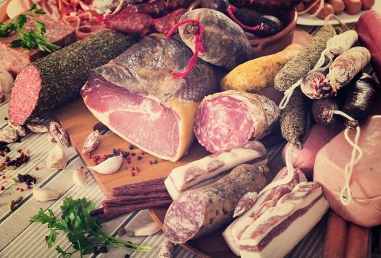 Diversos embutidos sobre una tabla de madera: salami, jamón, salchichas, tocino y perejil.