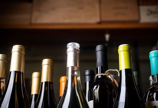 Botellas de vino con tapas revestidas de papel de aluminio de distintos colores, alineadas en un estante, con cajas de madera en el fondo.
