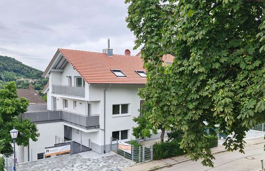 Weißes Gebäude mit rotem Dach, Balkonen und grünen Bäumen. Im Hintergrund ein Berg.