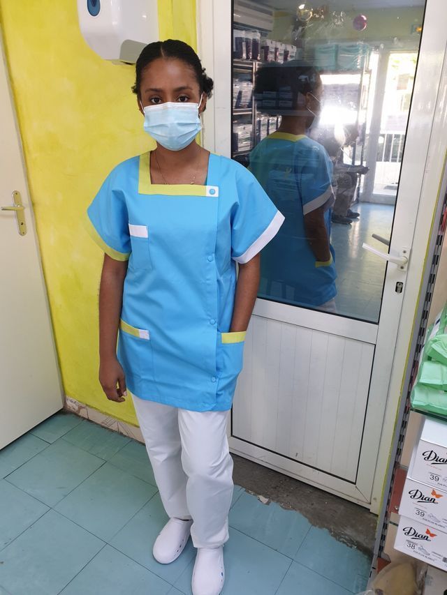 Une soignante en uniforme bleu et masque chirurgical se tient dans un couloir d'hôpital.