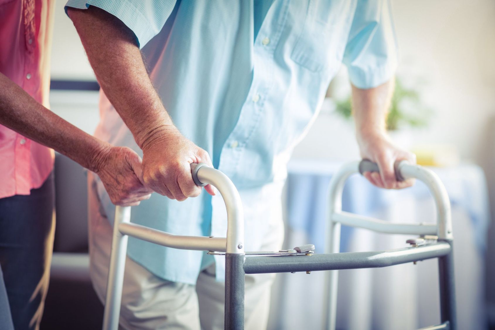 Une personne âgée en chemise bleue tient un déambulateur, et un aide-soignant se tient à ses côtés.