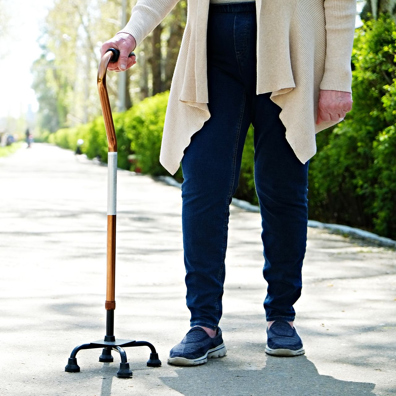 Une personne se tient debout avec une canne sur un trottoir ensoleillé, vêtue d'un jean, de baskets et d'un cardigan beige.